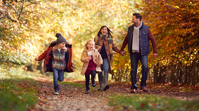 A family of four bundled up and walking through an autumnal, leafy woodland